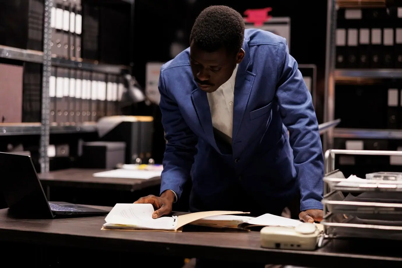 Афроамериканский менеджер работает над бухгалтерским отчетом, анализируя административные файлы в корпоративном хранилище. Бизнесмен проверяет бюрократический документ, ищет исследования по менеджменту.